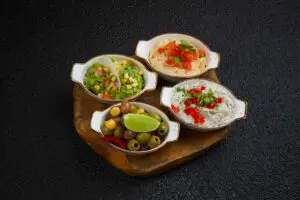 "An assortment of dips and olives served in small bowls on a wooden board. The platter includes guacamole with diced vegetables, hummus topped with diced tomatoes and herbs, a creamy dip garnished with red peppers and herbs, and a bowl of mixed olives with a lime wedge."