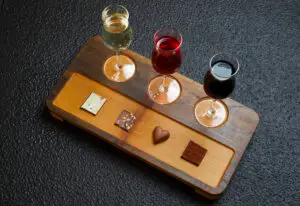 "Four glasses of red wine and an assortment of chocolates arranged on a wooden board, each chocolate piece placed in front of its corresponding wine glass."